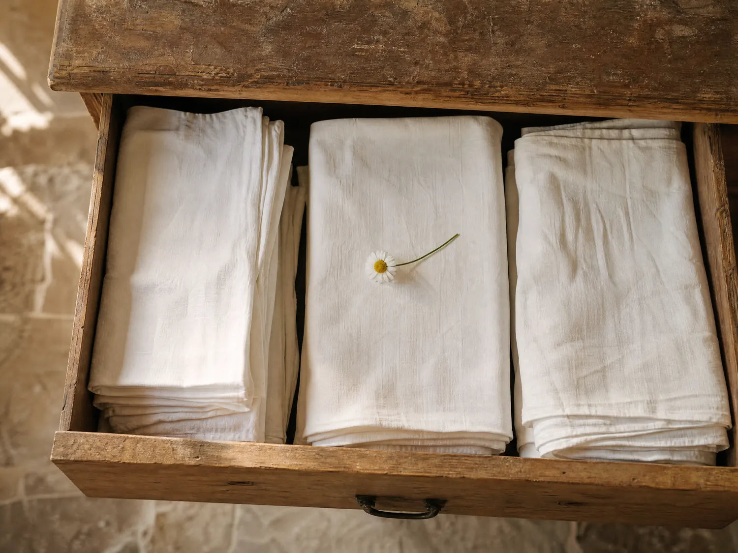 Il cassetto aperto di un vecchio cassettone di legno, con tre pile di biancheria di lino bianca piegata e una margherita di campo appoggiata sopra.