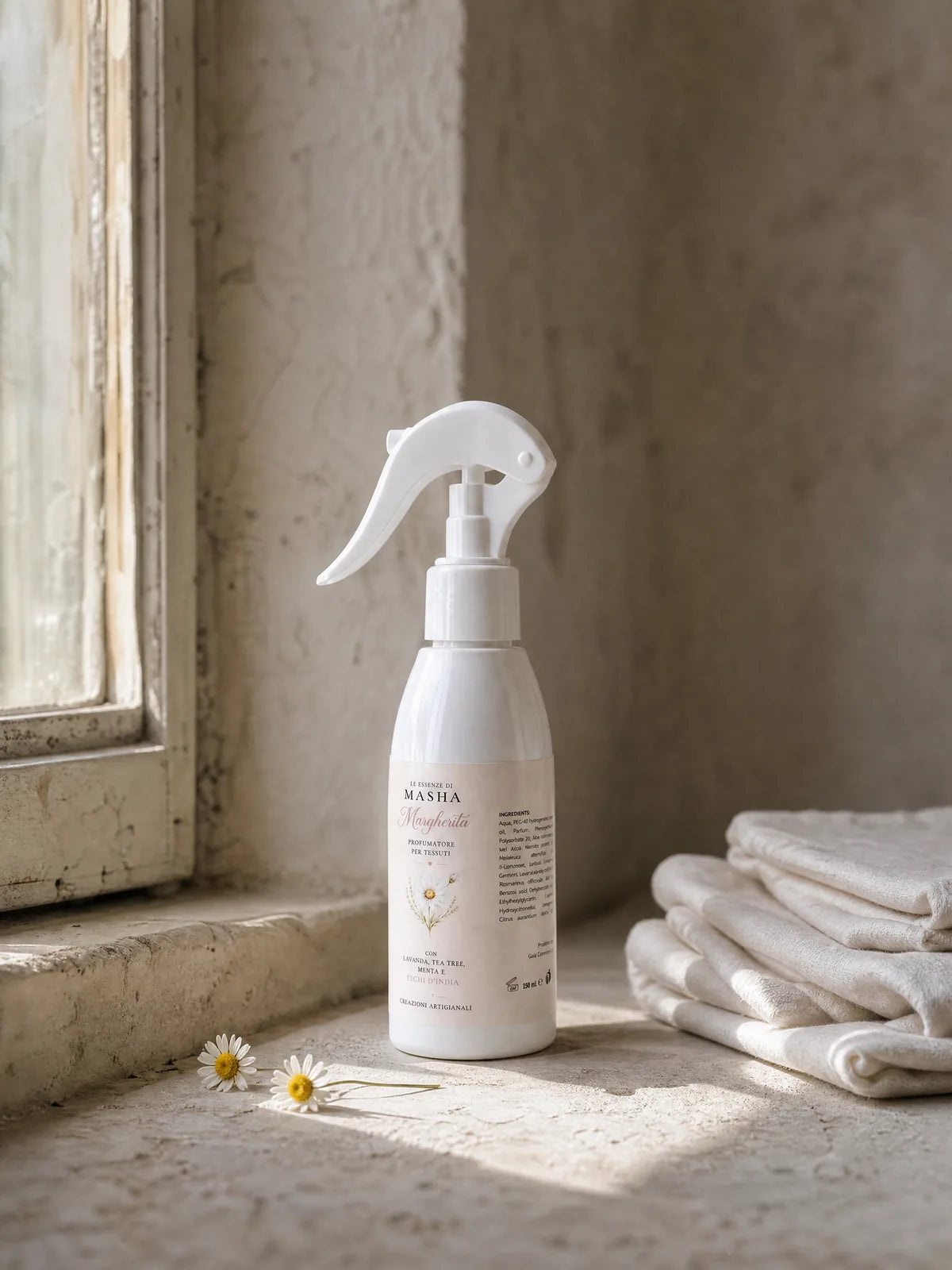 Il flacone spray Margherita da 150 ml, con la sua etichetta color crema, su un davanzale di pietra in una stanza dai muri a calce, accanto a una pila di lino piegato e due margherite di campo.