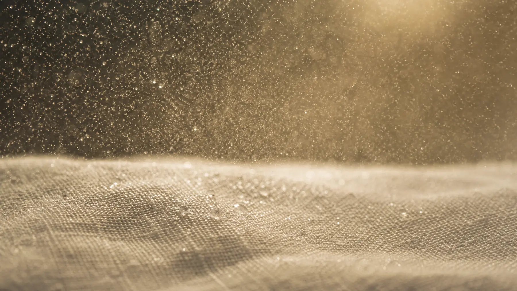 Macro in controluce: la nebbia finissima dello spray sospesa nell'aria sopra un tessuto di lino, le goccioline che prendono la luce.